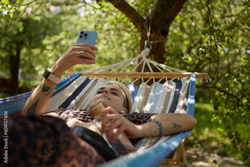 Young diverse woman with short hair lying on a hammock in a park and browsing mobile app on a smartphone. Stylish tattooed female relaxing in a green garden on weekend in summer