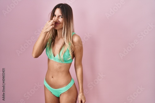 Young hispanic woman wearing bikini over pink background smelling something stinky and disgusting, intolerable smell, holding breath with fingers on nose. bad smell