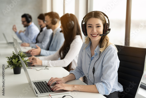 Friendly telephone operator woman with headset having conversation with client, showing excellent communication skills and attentive customer service, surrounded by group of colleagues at call center.