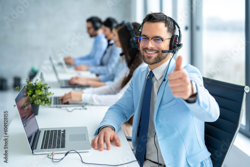 Male customer service representative showing approval sign with thumbs up while working in a row with his other operators at call center office