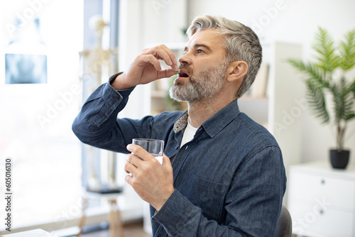 Caucasian middle-aged caucasian man taking pill and drinking glass of fresh water in doctor's office. Grey-bearded male person dressed in denim shirt following cardiologist's instructions.