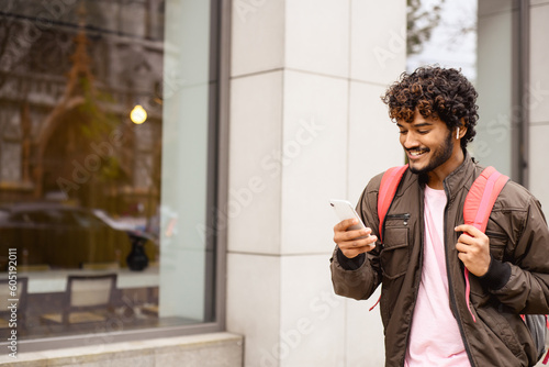 Positive indian man with backpack using smartphone and wireless earphones on urban street 