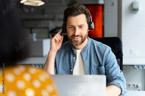 Handsome smiling call center operator using laptop, answering call, communication with customer sitting in modern office. Freelancer having video call at workplace