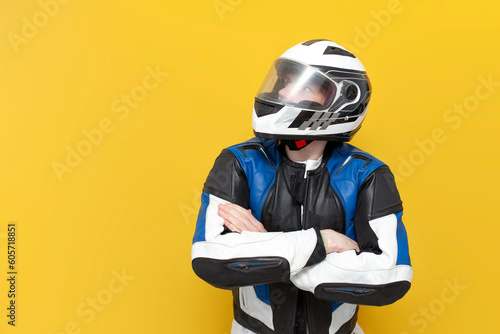 young guy motorcyclist in leather jacket and helmet stands with his arms crossed on yellow isolated background