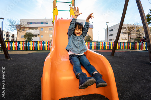 A cute Dominican girl is playing on a swing on a playground. The little girl is going down the slide happily. Concept of dark-skinned girls playing in a park, leisure among boys, playgrounds.