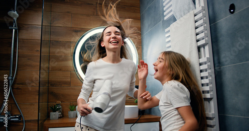 Funny young woman and 10 years old girl with long hair in pajamas drying her voluminous hair by hair dryer while dancing laughing and looking at each other in modern bathroom in home