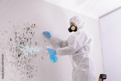Woman in protective suit and rubber gloves spraying mold remover onto wall