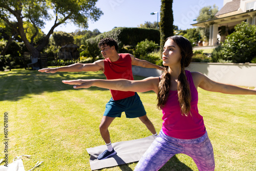 Biracial young couple practicing warrior 2 pose while standing on mat over grassy field in yard