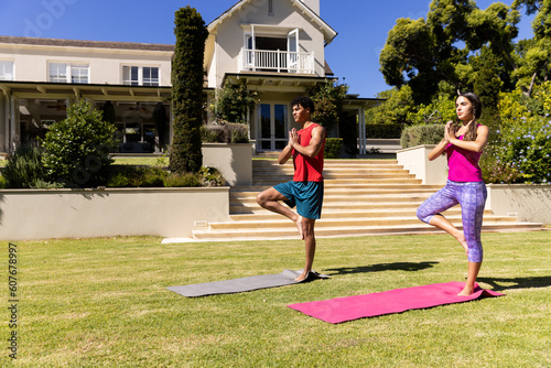 Biracial young couple standing on one leg and practicing meditation in yard against house