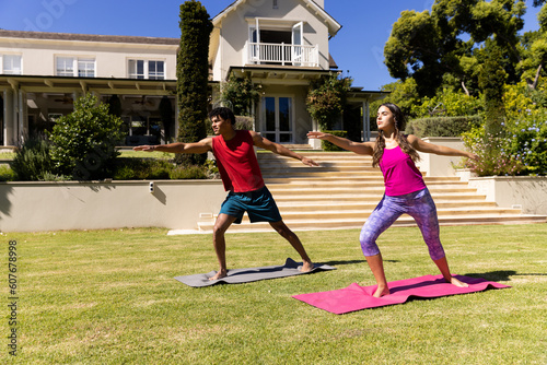 Biracial young couple practicing warrior 2 pose while standing on mats in yard against house
