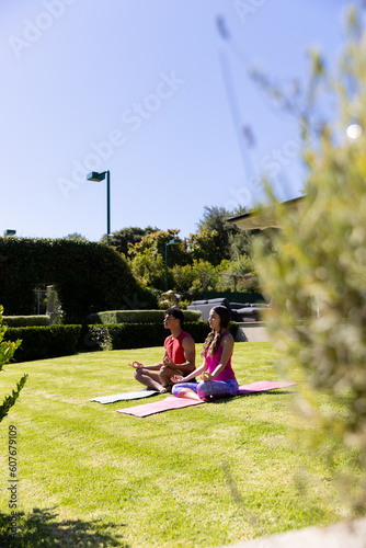 Biracial young couple practicing meditation while sitting on mats over field in yard under clear sky