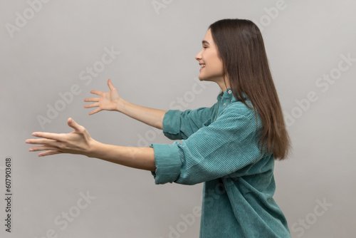 Come into my arms, free hugs. Side view of adorable hospitable woman smiling and reaching out hands, going to embrace, wearing casual style jacket. Indoor studio shot isolated on gray background.