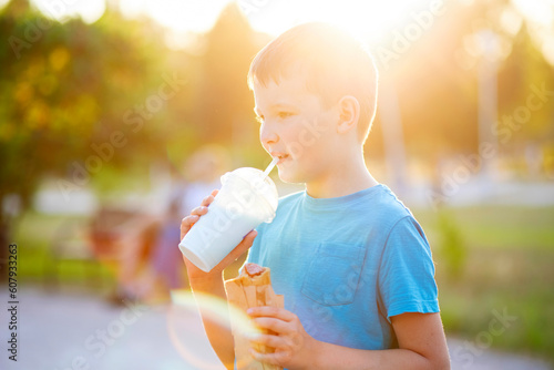 Child boy eats hot-dog sandwich and drinks milkshake the park at sunset outdoors