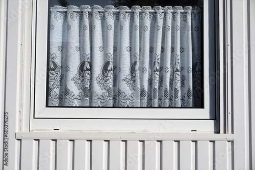 Fenster mit Gardinenstange und weißen Gardinen mit grauem Muster von weißer Metallfassade einer Containers bei Sonne am Abend im Frühling