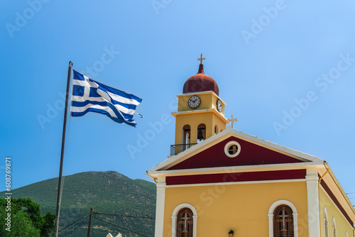 A colorful Christian church facade with a bell tower and a Greek flag waving against the blue sky on the Ionian Island of Cephalonia Greece.