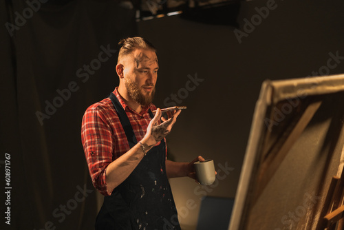 Male artist standing in his modern creative studio with large canvas, holds phone talks on speakerphone with friend, makes voice recognition or request uses virtual assistant