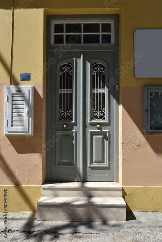 Old building face wall with entry door last repainted gray. Double doors with transom window and decorative metal grille. House entrance with marble steps leading to sunny street in Athens, Greece.