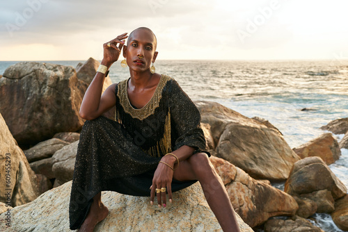 Non-binary ethnic fashion model in dress, brass jewelry sits on rocks by ocean . Trans sexual black person with rings, nose-ring, bracelets, earrings in posh clothes poses in tropical seaside location