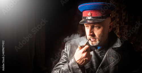 Officer from army of USSR. Man in NKVD cap. Investigator smokes pipe. KGB officer in leather coat. Symbol of USSR on cap. Member of soviet army. Portrait of man officer of KGB of USSR