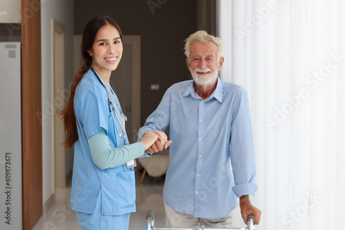 Happy caregiver helping senior old man walking with walking frame stand up from bedroom at home or retirement house..Asian smiling nurse taking good help care and support of elder patient at house.