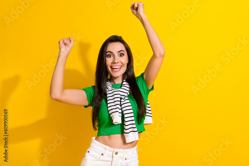 Photo of excited lucky girl dressed green t-shirt rising fists screaming empty space isolated yellow color background