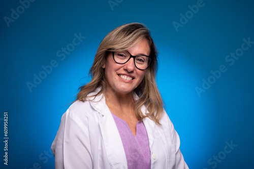 Photo of a sweet, confident mid-aged nurse dressed in a white coat and lilac blouse, with glasses looking at the camera.