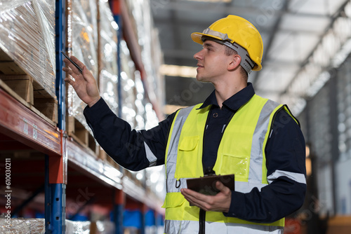 Male warehouse worker wearing uniform checks stock inventory in warehouse. Male worker holding clipboard and checking barcodes on boxes on shelf pallet in storage warehouse