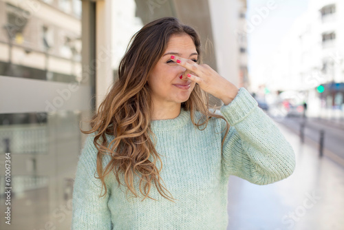 pretty hispanic woman feeling disgusted, holding nose to avoid smelling a foul and unpleasant stench