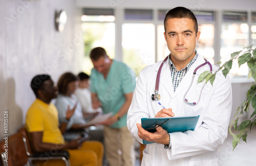 Portrait of focused man, professional doctor wearing white coat standing in lobby of clinic, filling out medical form at clipboard