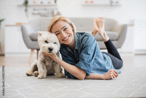 Full length portrait of lovely smiling woman in denim shirt caressing adorable little Westie on kitchen floor of apartment. Cheerful pet keeper posing with good-natured small animal in home interior.