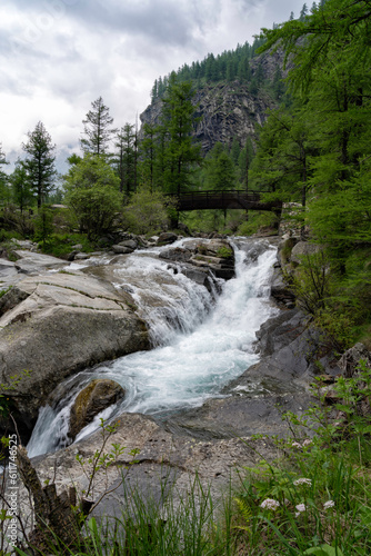 Mountain water stream and waterfall in the green forest with a wooden bridge on a cloudy day. Campiglia Soana (Azaria), Gran Paradiso National Park