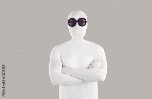 Portrait of person in bodysuit disguise and sunglasses. Man with secret, hidden personality, wearing white spandex costume and black round glasses standing with folded arms on grey color background