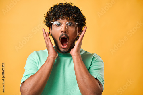 Close-up portrait of a handsome Indian man with curly hair in a turquoise t-shirt and glasses with big eyes and open mouth standing on a yellow background and expressing the emotion of shock, surprise