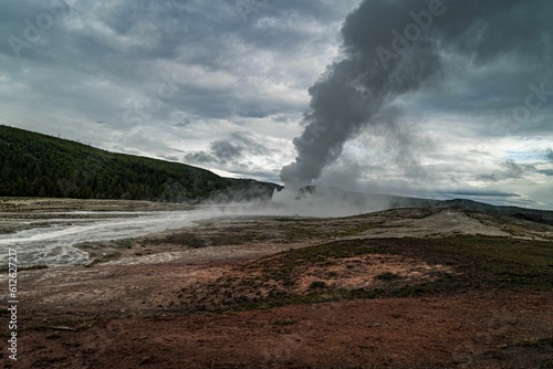 Scenic view of white smoke from a geyser with green mountain in the background on a cloudy day