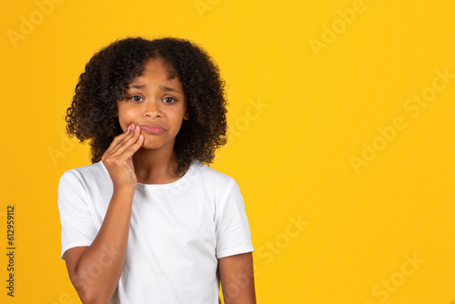 Sad curly teenager black schoolgirl in white t-shirt presses hand to cheek, suffering from toothache