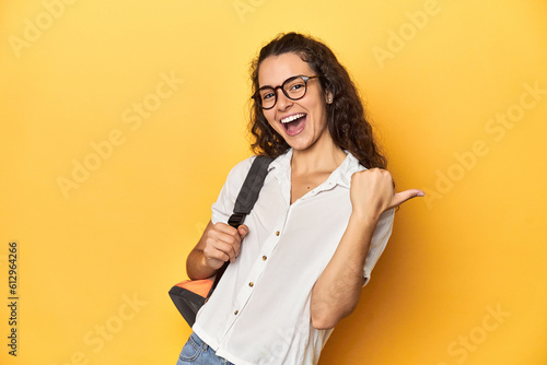 Caucasian university student with glasses, backpack, points with thumb finger away, laughing and carefree.