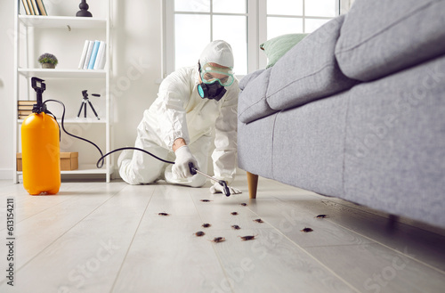 Pest control exterminator killing cockroaches inside house. Man in white protective suit, mask and gas respirator spraying insecticide from yellow sprayer bottle over roaches crawling on floor at home