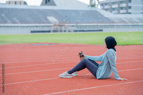 A young asian muslim woman wearing a black hijab is doing exercise and resting tired at the outdoor stadium in the morning. Modern Muslim woman concept, Muslim woman sport concept, Islam