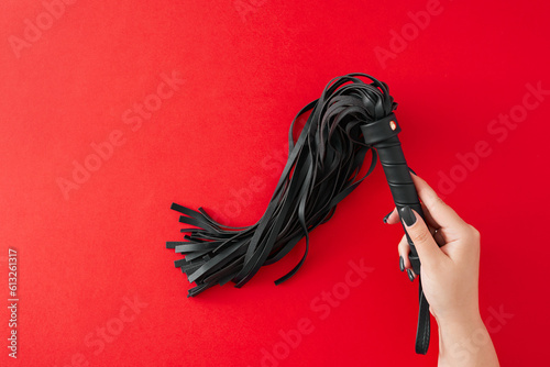 Ignite desires with carefully selected BDSM toys. First person top view photo of female hand holding leather whip on red background with empty space for text or advert