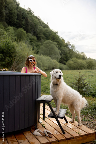 Woman bathing in outdoor hot tub while resting with her cute dog at house in mountains. Concept of recreation and spending leisure time with pets on nature