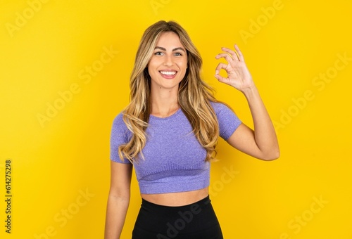 young beautiful blonde woman wearing sportswear over yellow studio background hold hand arm okey symbol toothy approve advising novelty news