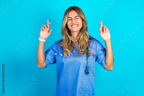 Young caucasian blonde doctor woman wearing blue uniform has big hope, crosses fingers, believes in good fortune, smiles broadly. People and wish concept