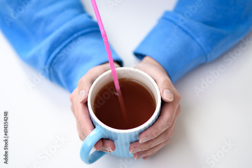 Kubek z gorącą herbatą trzymany w dłoniach, różowa plastikowa słomka jednorazowego użytku w środku 