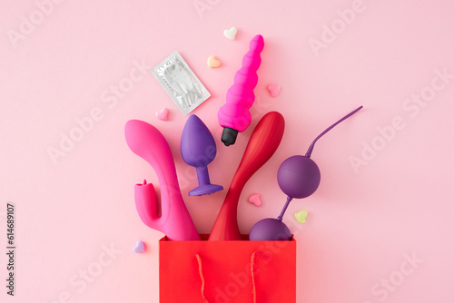 Concept of a store for sexual products. Top view flat lay of red shopping bag, vibrators, anal plugs, vaginal balls, condom, hearts on pastel pink background