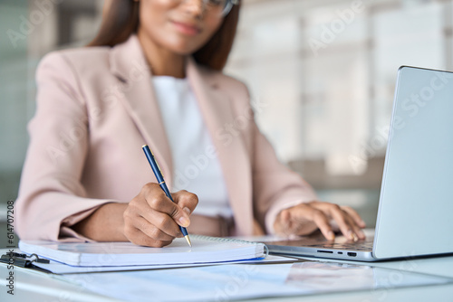 Close up view of professional busy African American business woman manager executive working on laptop computer writing notes in notebook at work elearning in corporate office.