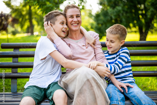 Happy mother is sitting with her sons on bench in park. They are having fun together.