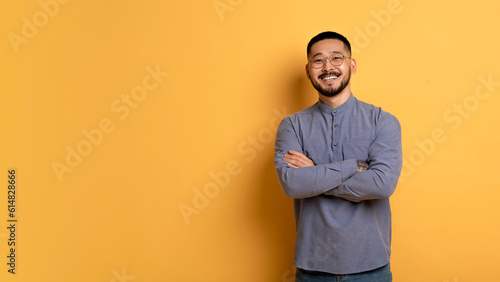 Confident young asian man standing with folded arms over yellow background