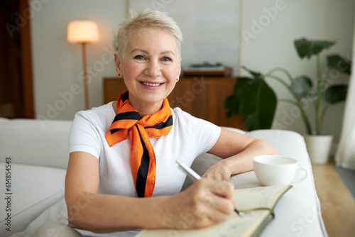 Portrait of cheerful happy female of 50s writing in her diary, planning next week, looking at camera with candid smile. Elderly woman making notes in her notepad sitting on couch in living-room