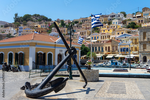 Anchor and flags of Greece on square of harbor town of Symi. Greek mountainous island and municipality, part of Dodecanese island chain. Adjacent upper town Ano Symi
