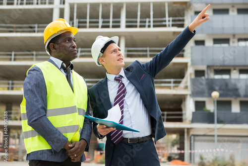 African-american foreman and Caucasian engineer in hardhats standing on construction site and talking. One man holding paper folder in hands, another one pointing with finger.